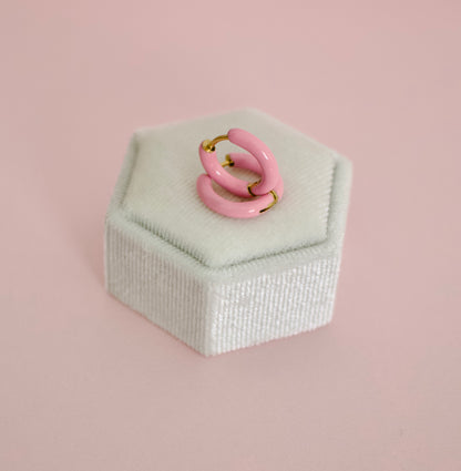 A pair of hypoallergenic pink enamel hoop earrings with gold-tone metal wire loops, displayed on a white velvet ring box against a solid pink background from the Pierced Peanut earring co.