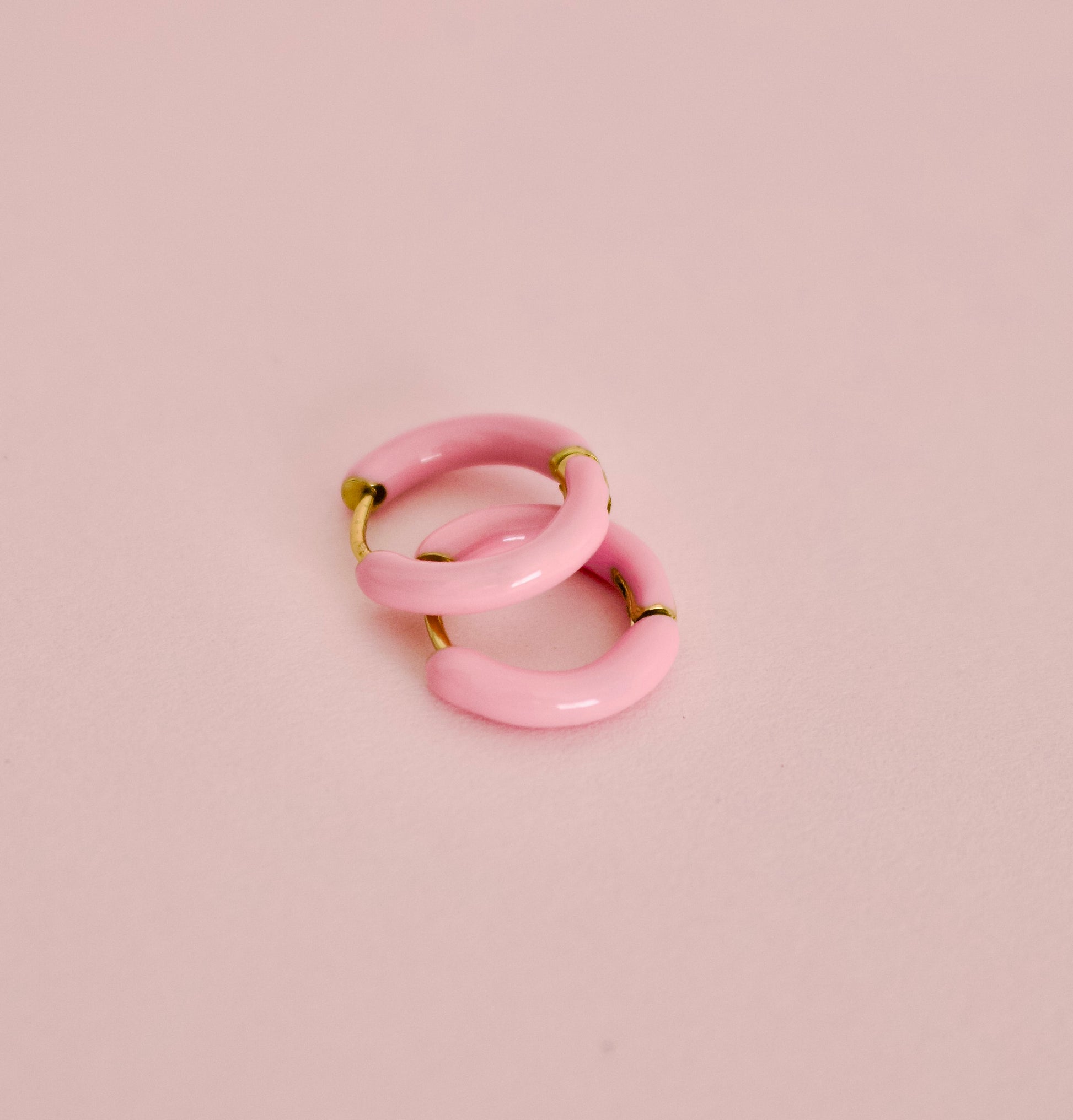 Two small hypoallergenic pink hoop earrings with gold-tone metal tips, arranged overlapping on a soft pink background from the Pierced Peanut.