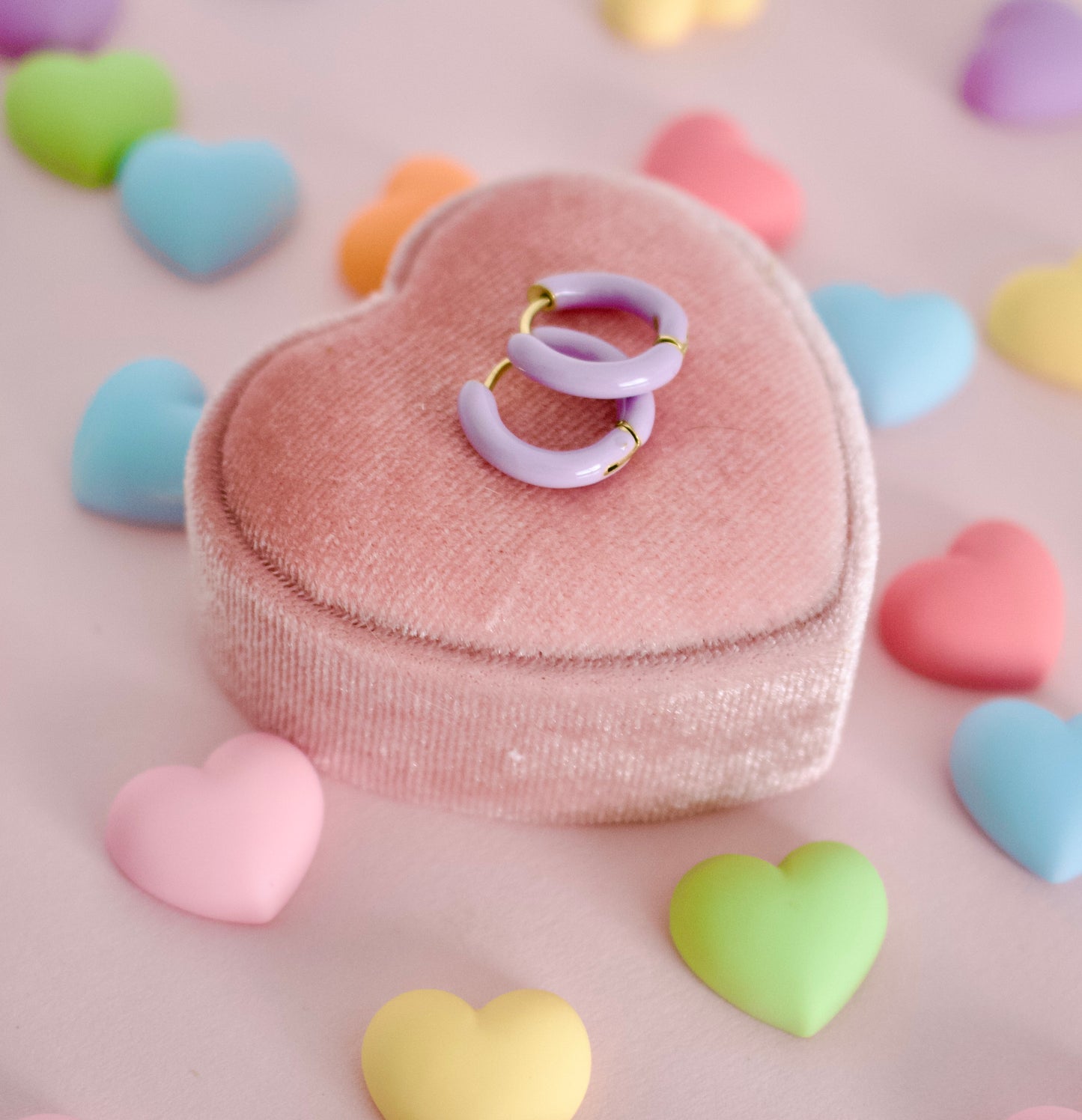 Two hypoallergenic pastel lavender hoop huggie earrings with gold-tone edges resting on a pink heart-shaped velvet jewelry box, surrounded by scattered pastel heart-shaped candies on a pink background. The earrings are from the Pierced Peanut.