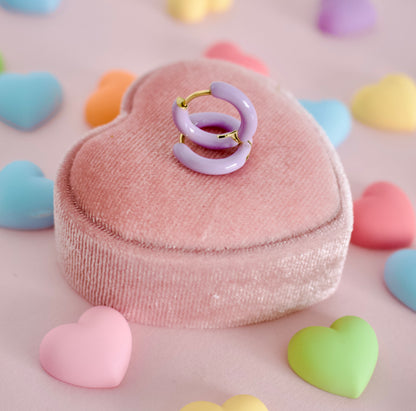 Two hypoallergenic lavender hoop huggie earrings with gold-tone rings, displayed on a pink heart-shaped velvet jewelry box surrounded by pastel heart-shaped candies on a pink background. The earrings are from the kids earring brand the Pierced Peanut.
