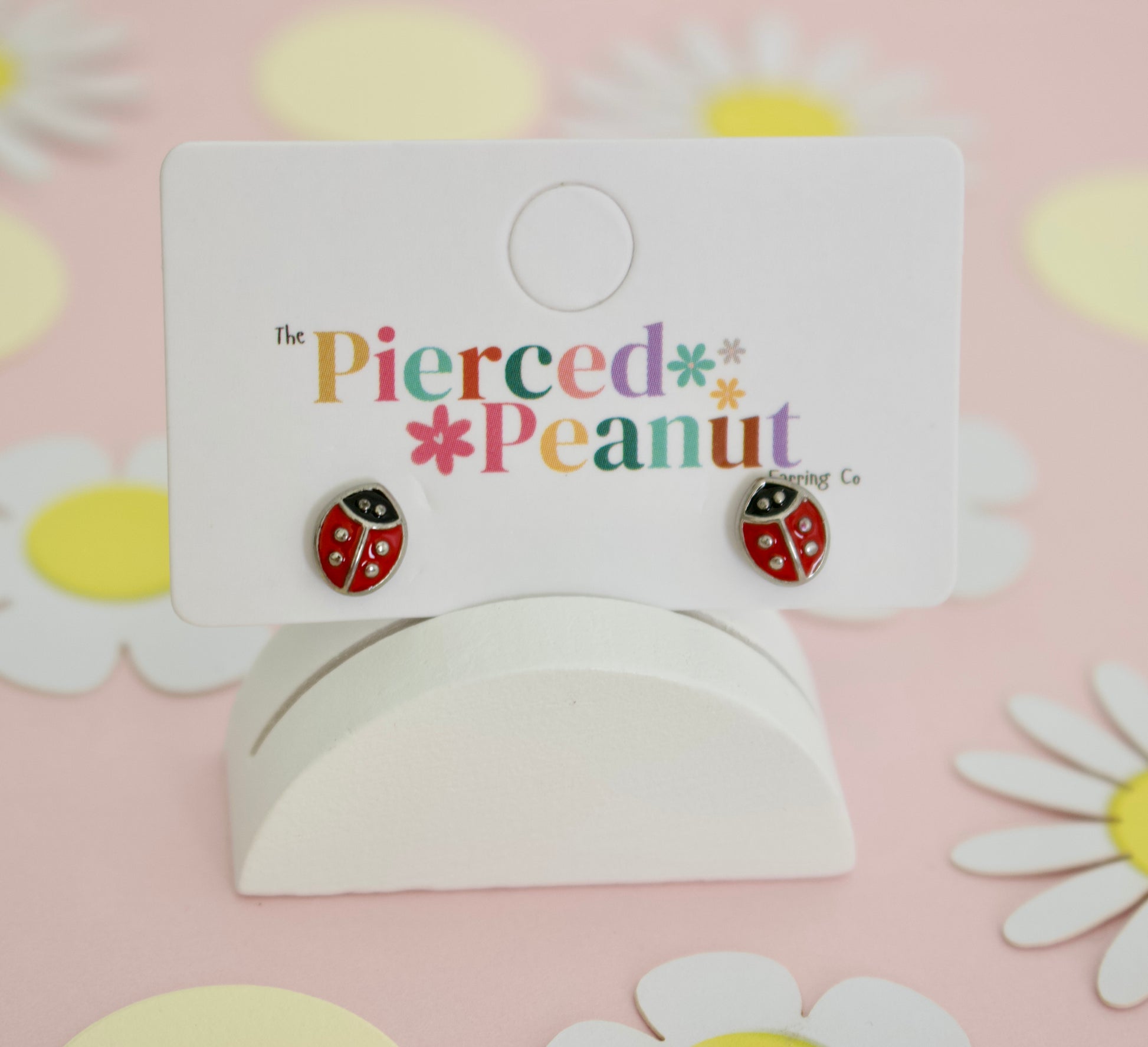 A pair of hypoallergenic gold toned ladybug screwback stud earrings with red enamel details. The kids earrings are displayed on a branded display card and are from the childrens brand the Pierced Peanut earring company.