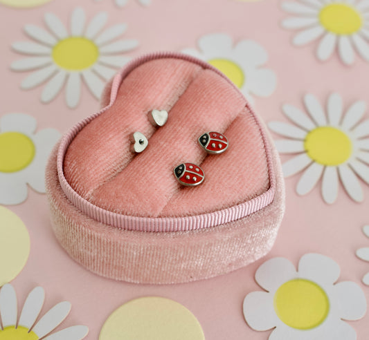A pair of hypoallergenic gold toned ladybug screwback stud earrings with red enamel details. The kids earrings are displayed on a pink heart shaped box and are from the childrens brand the Pierced Peanut earring company.