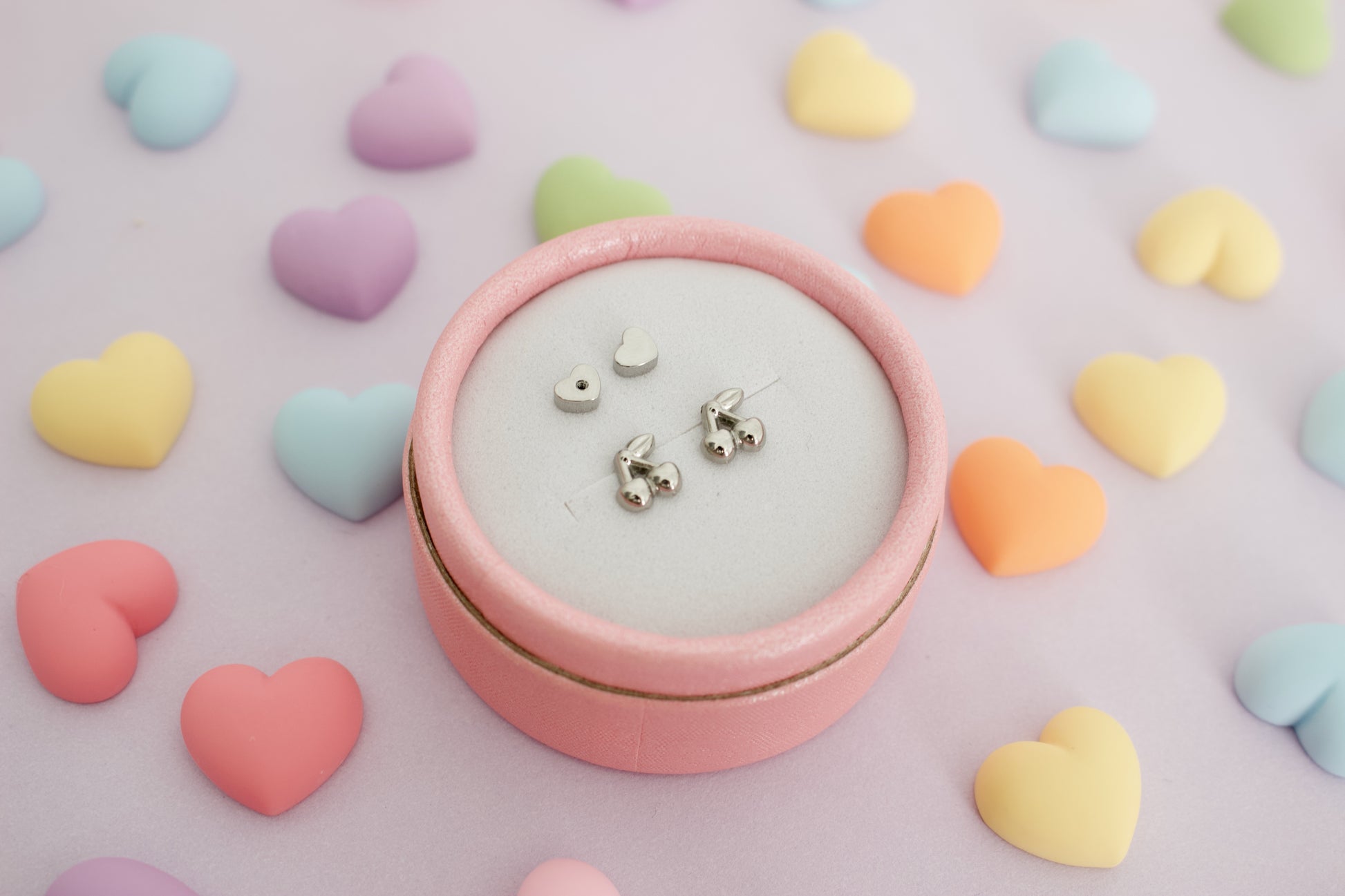 A pair of hypoallergenic kids silver cherry screwback stud earrings from The Pierced Peanut Earring Co with small heart-shaped backing pieces, arranged on a light pink surface surrounded by pastel heart-shaped candies.