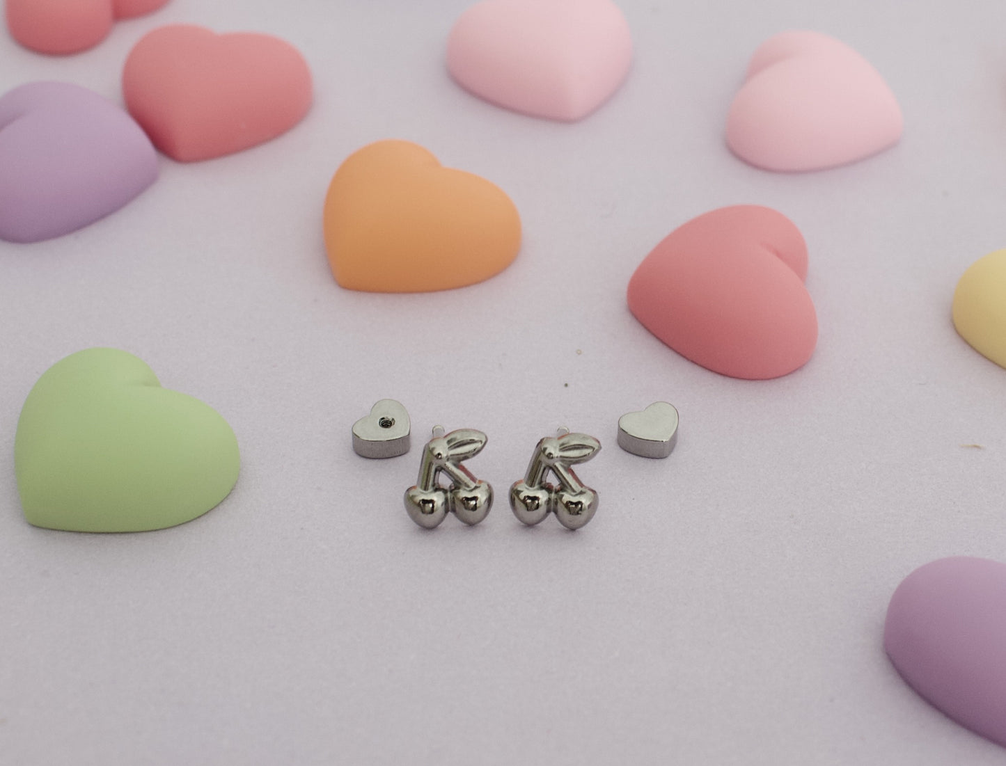 A pair of hypoallergenic kids silver cherry screwback stud earrings from The Pierced Peanut Earring Co with small heart-shaped backing pieces, arranged on a light pink surface surrounded by pastel heart-shaped candies.
