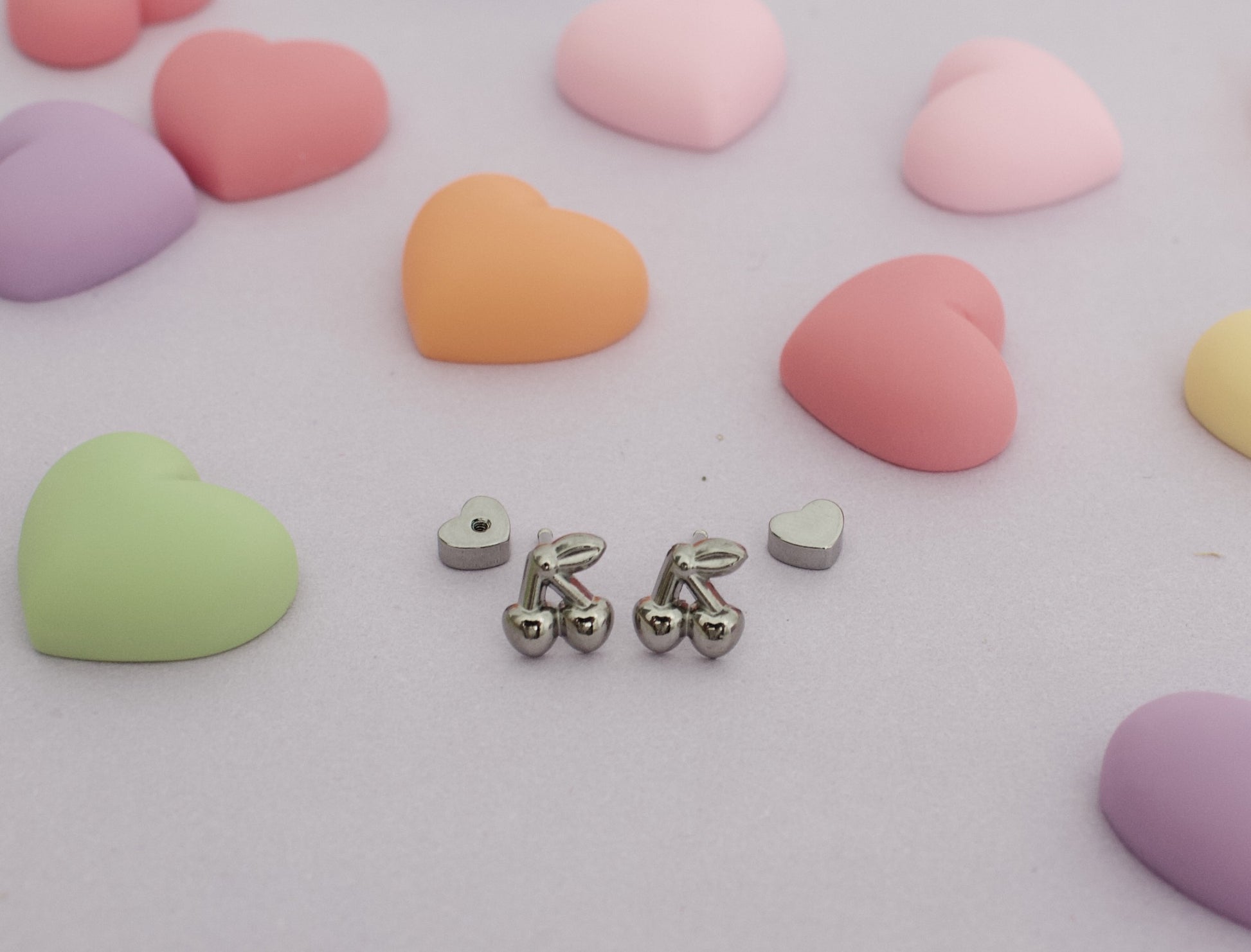 A pair of hypoallergenic kids silver cherry screwback stud earrings from The Pierced Peanut Earring Co with small heart-shaped backing pieces, arranged on a light pink surface surrounded by pastel heart-shaped candies.