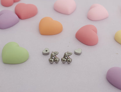 A pair of hypoallergenic kids silver cherry screwback stud earrings from The Pierced Peanut Earring Co with small heart-shaped backing pieces, arranged on a light pink surface surrounded by pastel heart-shaped candies.