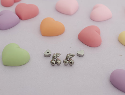 A pair of hypoallergenic kids silver cherry screwback stud earrings from The Pierced Peanut Earring Co with small heart-shaped backing pieces, arranged on a light pink surface surrounded by pastel heart-shaped candies.