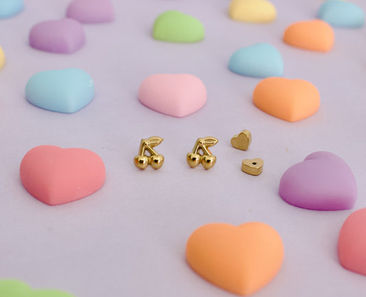 Gold cherry hypoallergenic screwback stud earrings for kids from The Pierced Peanut Earring Co. They have a small heart-shaped back, displayed on a pastel lavender background surrounded by colorful candy heart props.
