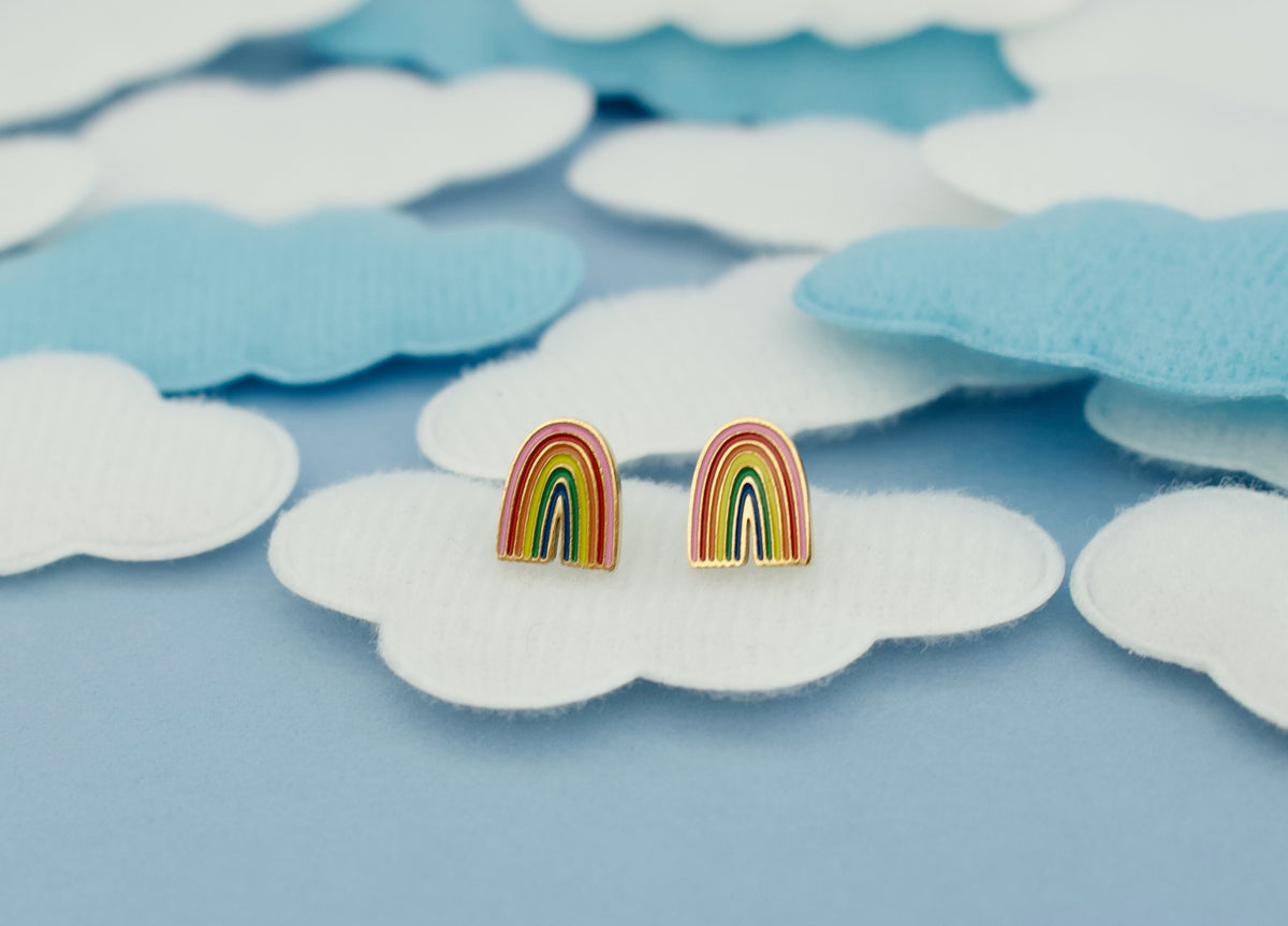 A pair of kids hypoallergenic rainbow stud earrings with gold-toned posts and heart shaped screw backs, displayed on a light blue background with white and light blue cloud-shaped fabric pieces from the Pierced Peanut.
