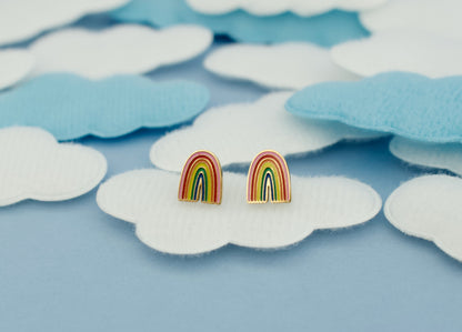 A pair of kids hypoallergenic rainbow stud earrings with gold-toned posts and heart shaped screw backs, displayed on a light blue background with white and light blue cloud-shaped fabric pieces from the Pierced Peanut.