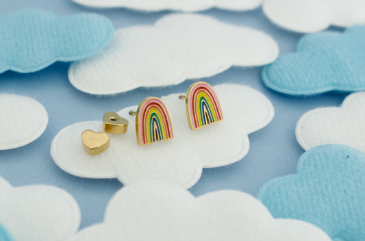 A pair of kids hypoallergenic rainbow stud earrings with gold-toned posts and heart shaped screw backs, displayed on a light blue background with white and light blue cloud-shaped fabric pieces from the Pierced Peanut.