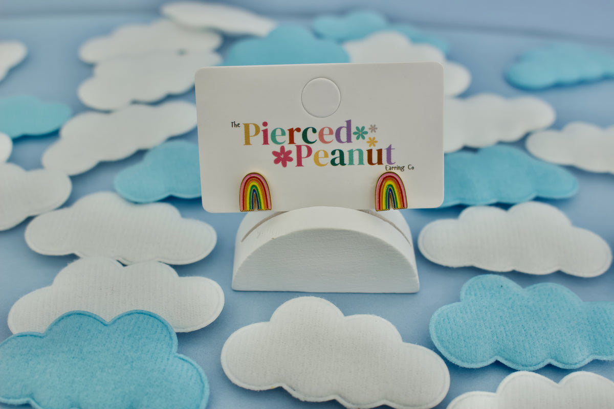 A pair of kids hypoallergenic screwback rainbow stud earrings displayed on a white card stand. The card reads 'The Pierced Peanut Earring Co' and is set against a background of light blue and white felt cloud cutouts on a light blue surface.