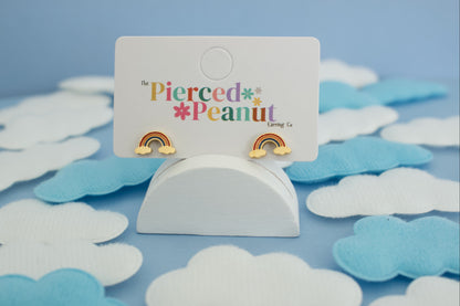 A pair of kids hypoallergenic gold rainbow stud earrings displayed on a white card labeled 'The Pierced Peanut Earring Co', set in a styled flat-lay with white and light blue cloud cutouts on a blue background.