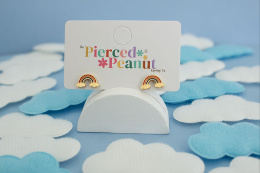 A pair of kids hypoallergenic gold rainbow stud earrings displayed on a white card labeled 'The Pierced Peanut Earring Co', set in a styled flat-lay with white and light blue cloud cutouts on a blue background.