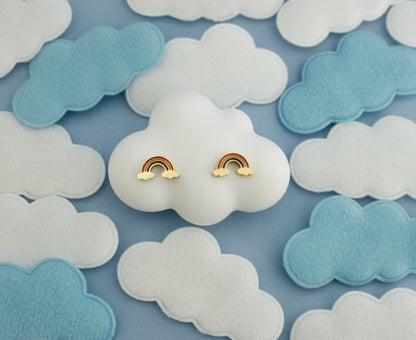 A pair of small kids hypoallergenic rainbow stud earrings with gold-tone metal and pastel rainbow enamel, displayed on a white cloud-shaped stand against a blue background with white and light blue felt clouds.