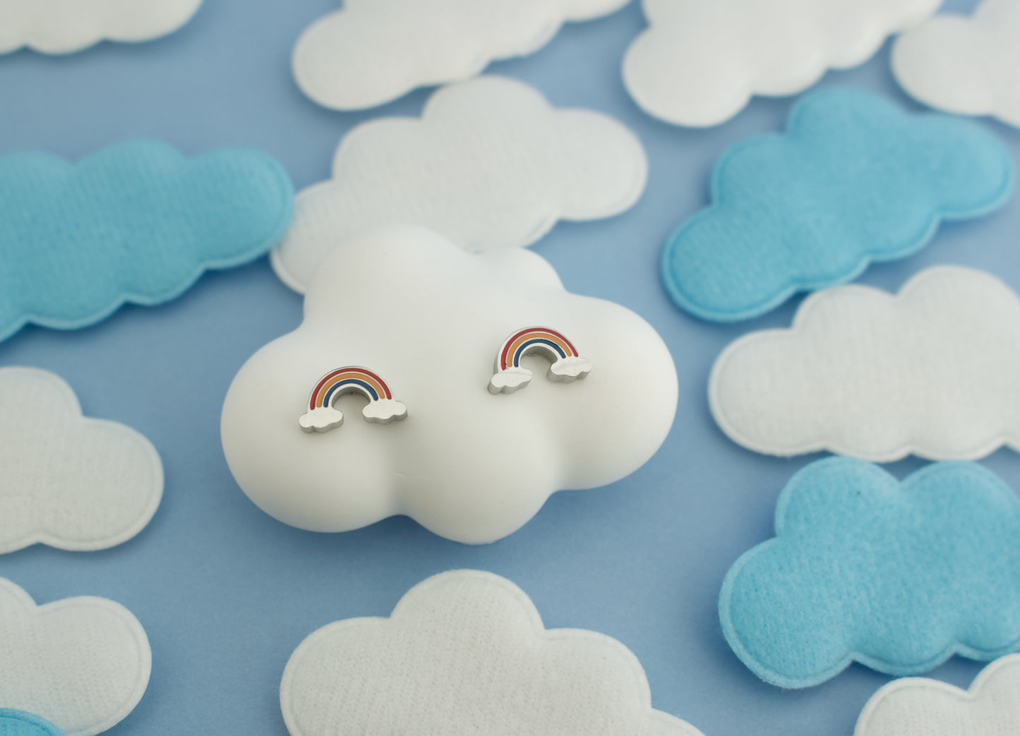 A pair of small kids hypoallergenic rainbow cloud screwback stud earrings displayed on a white cloud-shaped stand, surrounded by white and light blue felt cloud decorations on a light blue background.
