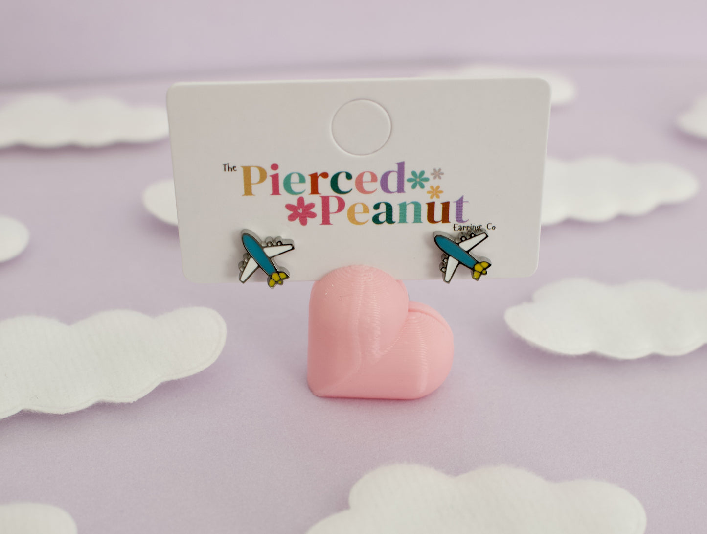 A pair of kids small hypoallergenic airplane screwback stud earrings displayed on a white branded card, with a pink heart-shaped stand in front. The card reads 'The Pierced Peanut,' and the background is a soft lavender surface with white cloud cutouts.