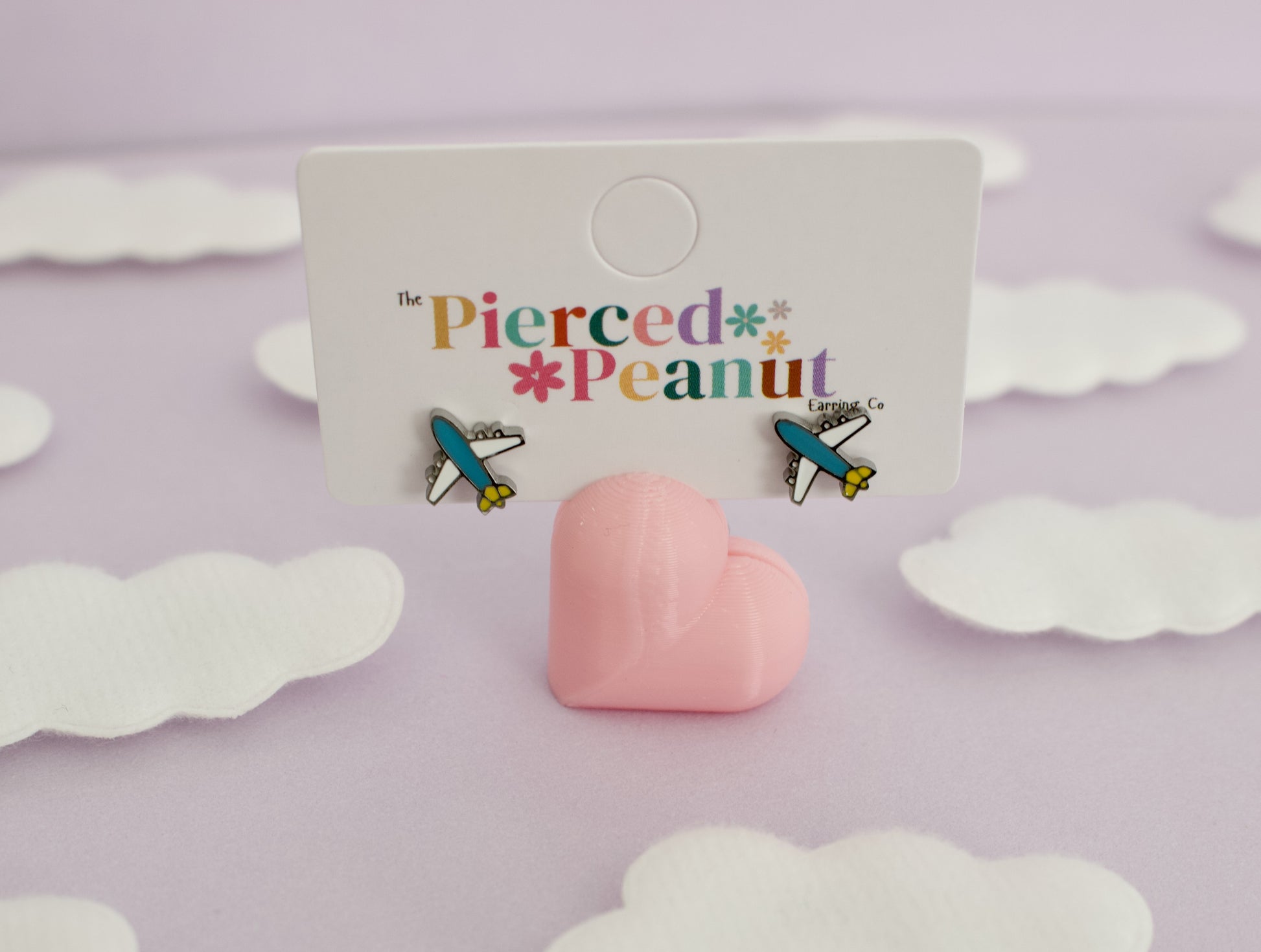 A pair of kids small hypoallergenic airplane screwback stud earrings displayed on a white branded card, with a pink heart-shaped stand in front. The card reads 'The Pierced Peanut,' and the background is a soft lavender surface with white cloud cutouts.