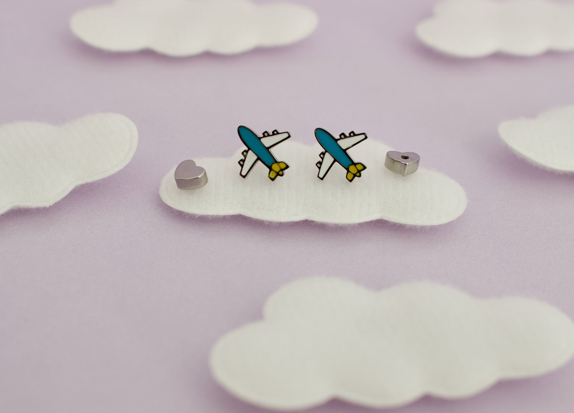 A pair of small kids hypoallergenic airplane screwback stud earrings displayed on a white cloud-shaped backing against a soft lavender background with additional cloud cutouts.