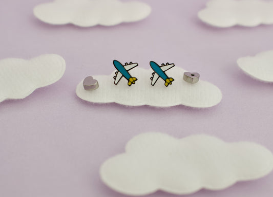 A pair of small kids hypoallergenic airplane screwback stud earrings displayed on a white cloud-shaped backing against a soft lavender background with additional cloud cutouts.