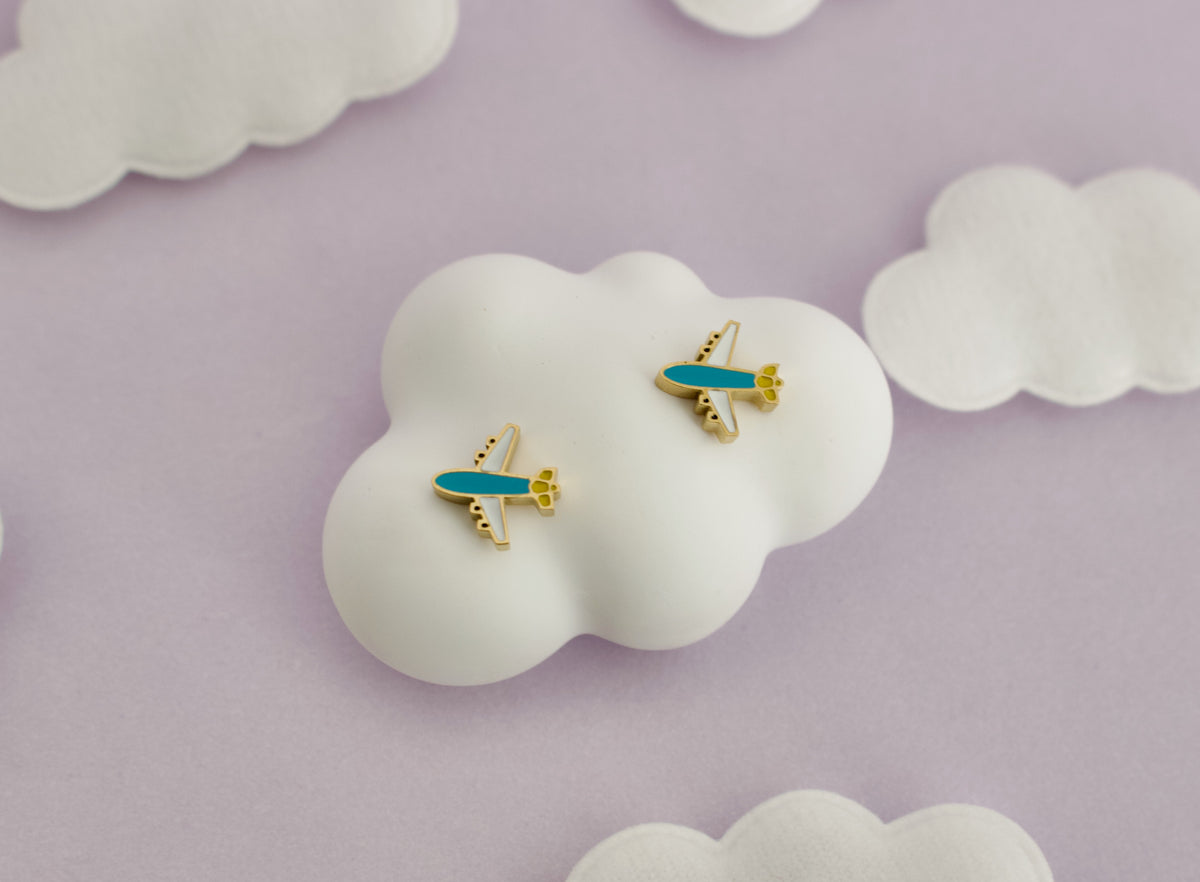 A pair of small kids hypoallergenic airplane screwback stud earrings with turquoise blue bodies and gold accents, displayed on a white cloud-shaped stand against a soft lavender background with additional white cloud props.