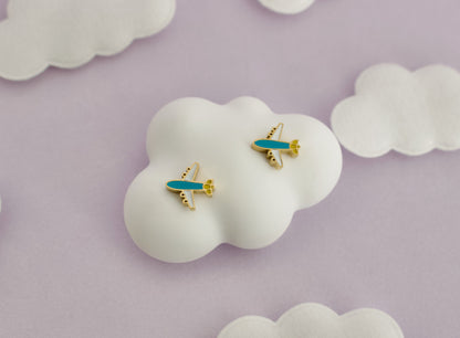 A pair of small kids hypoallergenic airplane screwback stud earrings with turquoise blue bodies and gold accents, displayed on a white cloud-shaped stand against a soft lavender background with additional white cloud props.