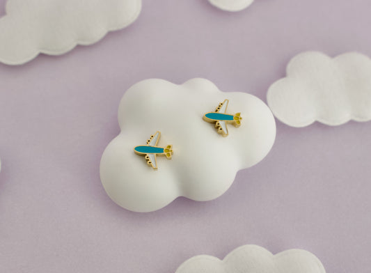 A pair of small kids hypoallergenic airplane screwback stud earrings with turquoise blue bodies and gold accents, displayed on a white cloud-shaped stand against a soft lavender background with additional white cloud props.