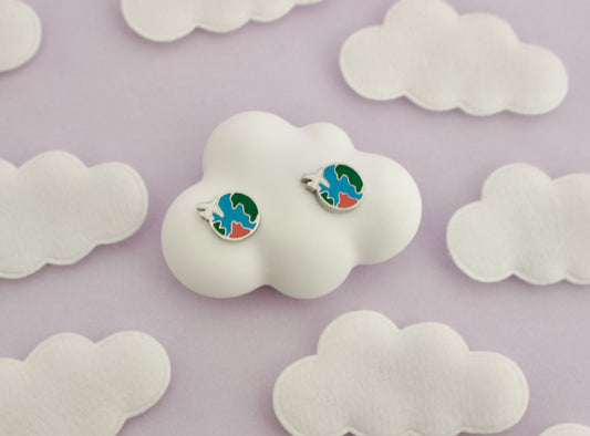 A pair of kids small hypoallergenic round silver screwback stud earrings with colorful enamel details, displayed on a white cloud-shaped stand against soft cloud cutouts from The pierced peanut earring co.
