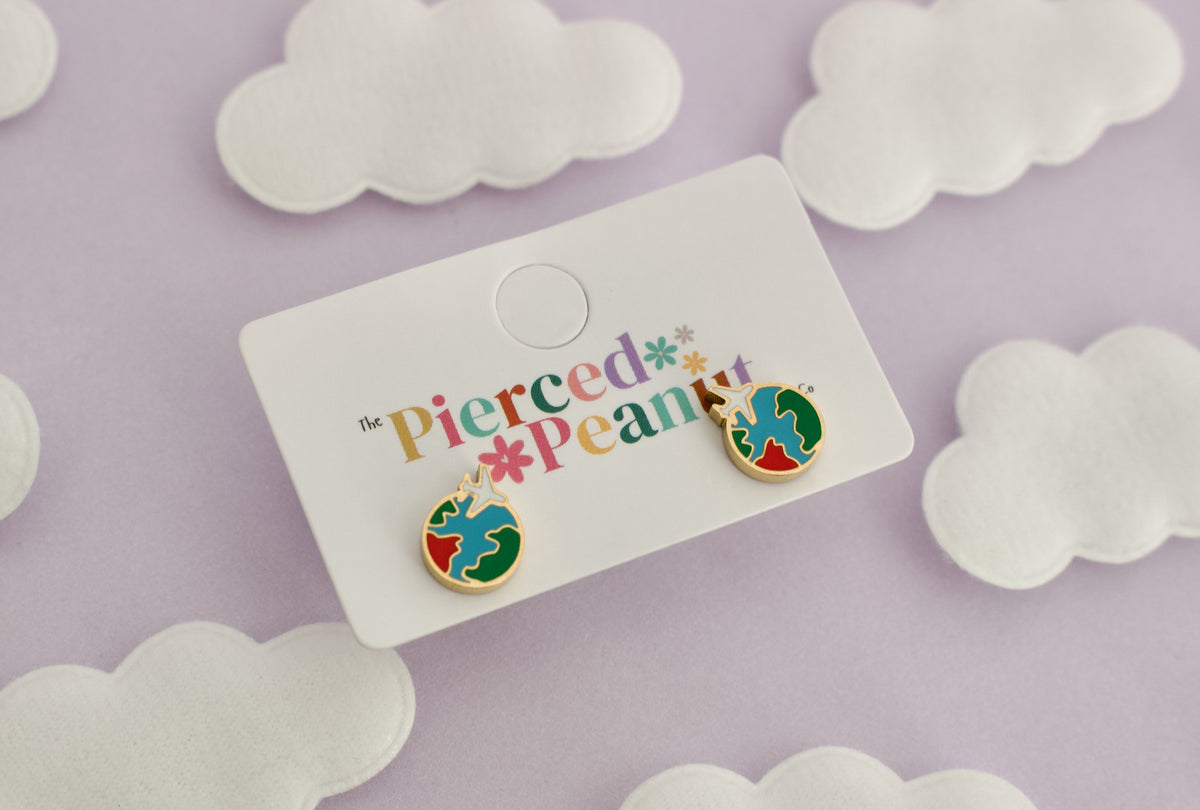 A pair of kids small hypoallergenic round gold-tone screwback stud earrings with colorful enamel details, displayed on a white cloud-shaped stand against a soft lavender background with white cloud cutouts from The Pierced Peanut Earring Co.