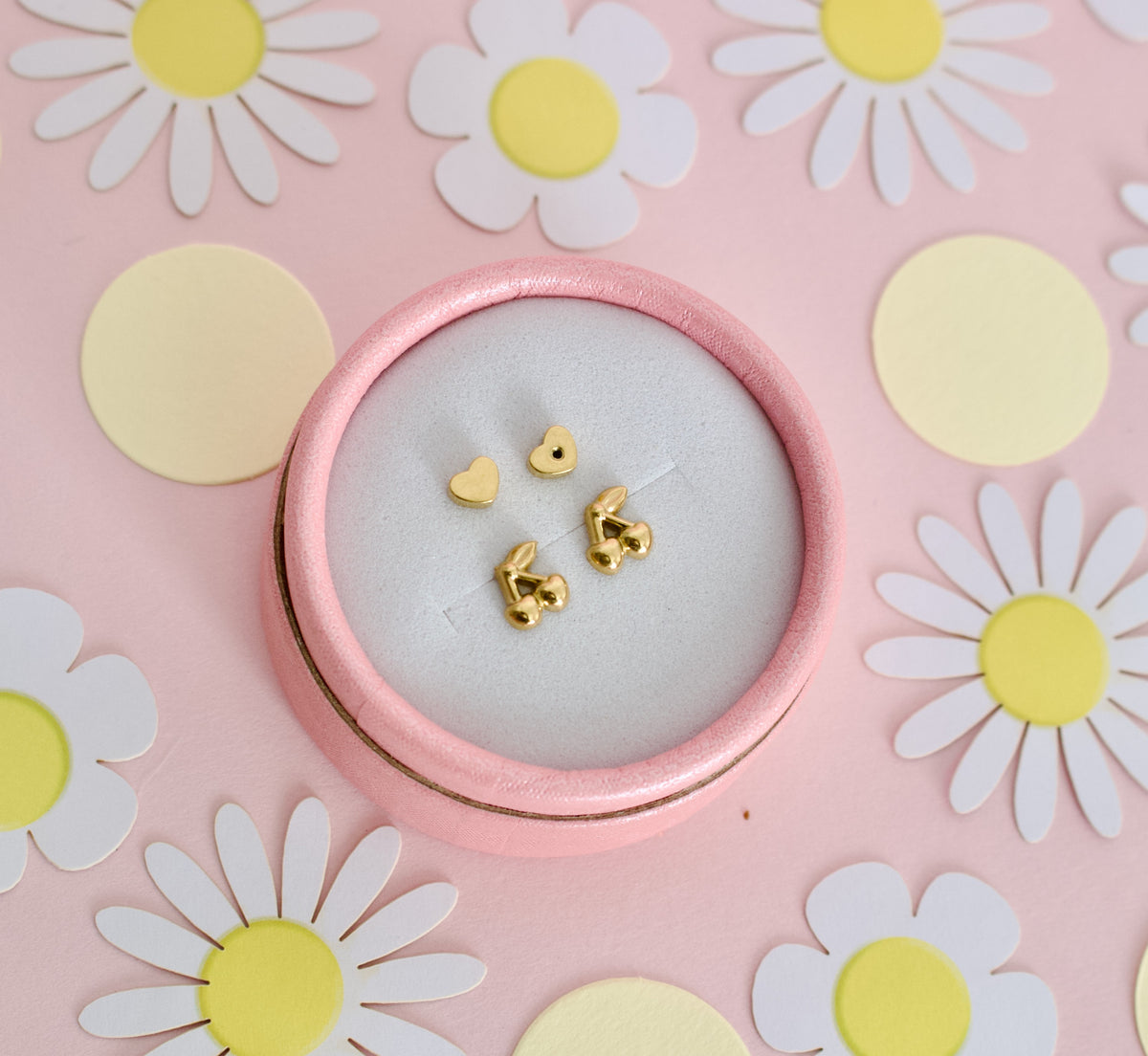 Gold cherry hypoallergenic screwback stud earrings for kids from The Pierced Peanut Earring Co. They have a small heart-shaped back, displayed on a pastel pink background surrounded by colorful candy flower props.
