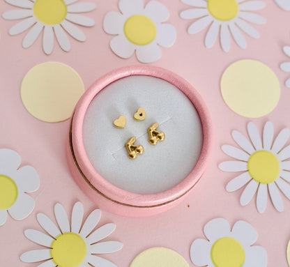 Gold cherry hypoallergenic screwback stud earrings for kids from The Pierced Peanut Earring Co. They have a small heart-shaped back, displayed on a pastel pink background surrounded by colorful candy flower props.