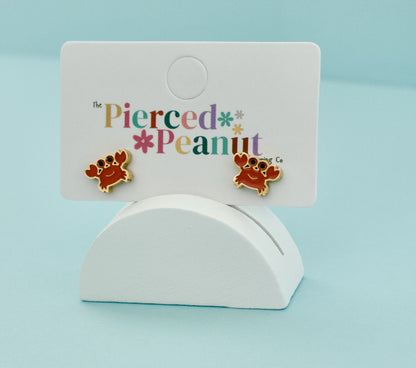 A pair of kids hypoallergenic, gold, crab screwback stud earrings with enamel orange details displayed on a jewellery card from The Pierced Peanut Earring Co.