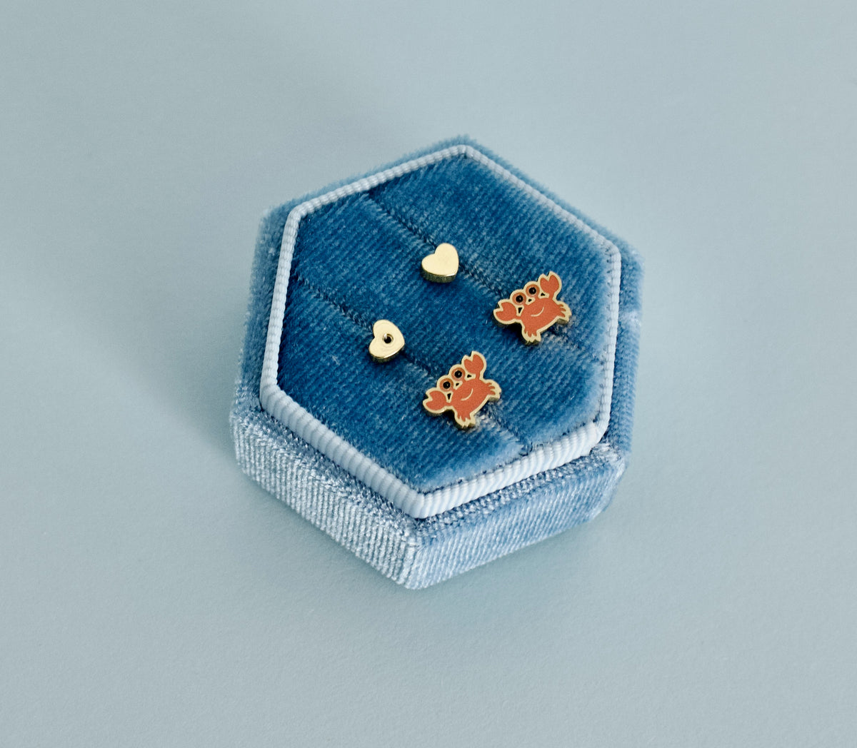 A pair of kids hypoallergenic, gold, crab screwback stud earrings with enamel orange details displayed in a blue jewellery box from The Pierced Peanut Earring Co.