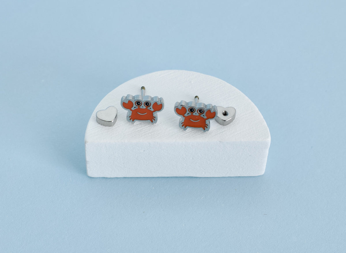 A pair of hypoallergenic kids silver-toned stud earrings with orange-red crab charms and gray claws, displayed on a white foam stand against a light blue background.
