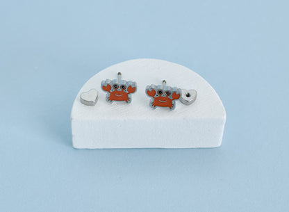 A pair of hypoallergenic kids silver-toned stud earrings with orange-red crab charms and gray claws, displayed on a white foam stand against a light blue background.