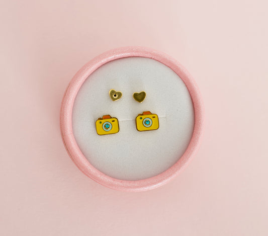 A pair of hypoallergenic kids yellow camera stud earrings with blue and orange details, displayed on a white circular backing inside a pink round box against a soft pink background. Earrings are from the brand The Pierced Peanut Earring Co in New Zealand.