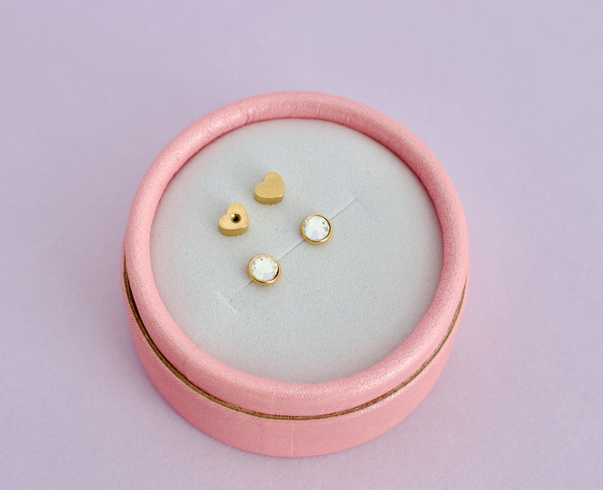 A Pair of hypoallergenic kids gold bezel cut diamond screwback studs displayed on a light gray velvet insert inside a round pink jewelry box against a solid lavender background. The brand is The Pierced Peanut Earring Co based in New Zealand.