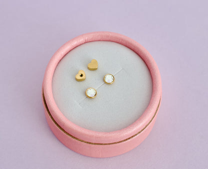 A Pair of hypoallergenic kids gold bezel cut diamond screwback studs displayed on a light gray velvet insert inside a round pink jewelry box against a solid lavender background. The brand is The Pierced Peanut Earring Co based in New Zealand.