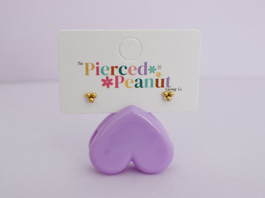A pair of kids small hypoallergenic gold stud earrings displayed on a white card labeled 'The Pierced Peanut Loving Co', with a lavender heart-shaped stand below on a pale purple background.