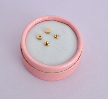 A pair of kids hypoallergenic gold trinity ball screwback stud earrings with heart shaped backs. Displayed inside a pale pink ring box from the brand The Pierced Peanut Earring Co based in New Zealand.