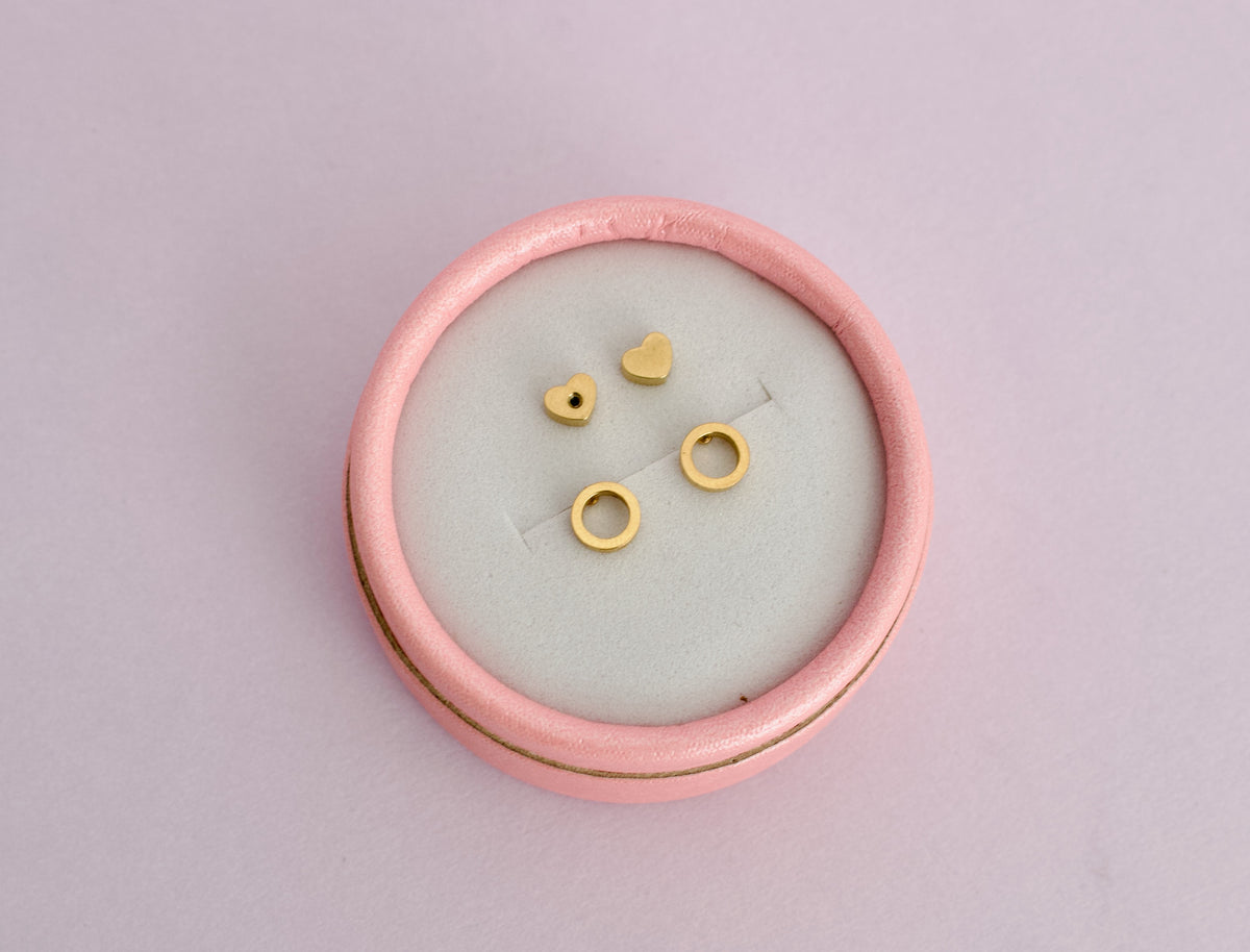 A pair of kids hypoallergenic gold open circle halo earrings. The studs have screwbacks in a heart shape and all are displayed on a pink box with a soft pink background from the Pierced Peanut Earring Co in Nz.