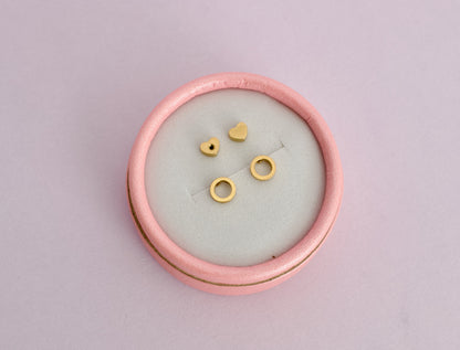 A pair of kids hypoallergenic gold open circle halo earrings. The studs have screwbacks in a heart shape and all are displayed on a pink box with a soft pink background from the Pierced Peanut Earring Co in Nz.