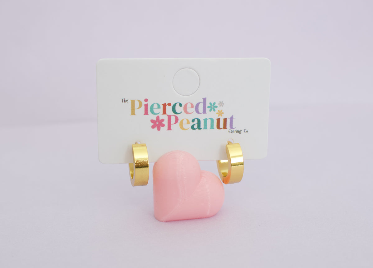 Kids hypoallergenic Gold hoop earrings on a small pink heart stand displayed on a white card from The Pierced Peanut Eending Co in New Zealand against a light lavender background.