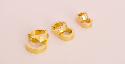 Three pairs of kids hypoallergenic shiny gold huggie hoop earrings arranged on a soft light pink background from the Pierced Peanut Earring Co in New Zealand, NZ.