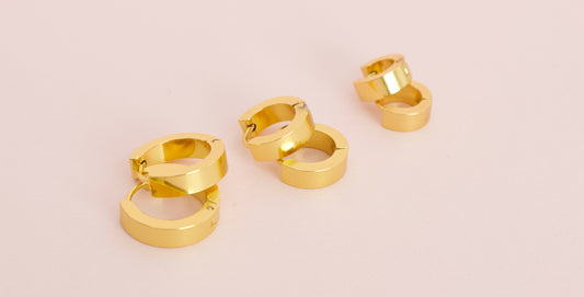 Three pairs of kids hypoallergenic shiny gold huggie hoop earrings arranged on a soft light pink background from the Pierced Peanut Earring Co in New Zealand, NZ.