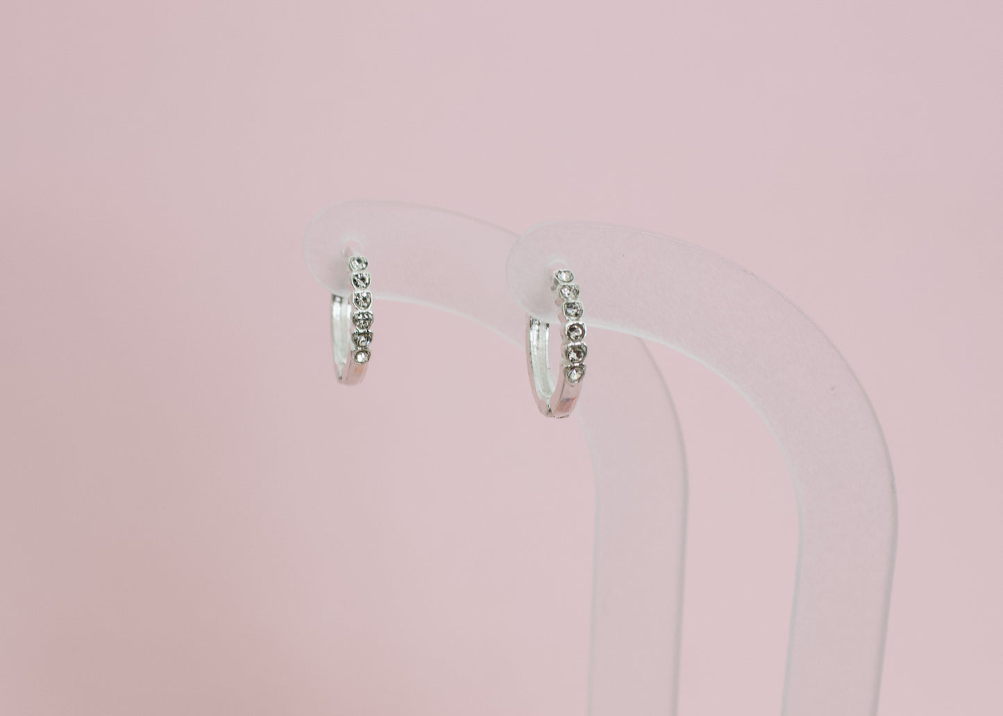 A pair of kids hypoallergenic silver earrings with small clear stones, displayed on a clear acryllic stand with a solid pink background. The earrings are from the brand The Pierced Peanut Earring Co in New Zealand, NZ.