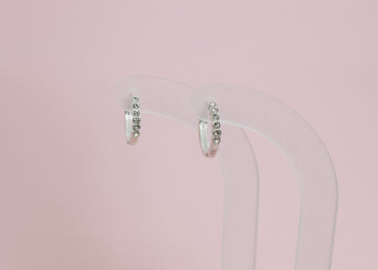 A pair of kids hypoallergenic silver earrings with small clear stones, displayed on a clear acryllic stand with a solid pink background. The earrings are from the brand The Pierced Peanut Earring Co in New Zealand, NZ.