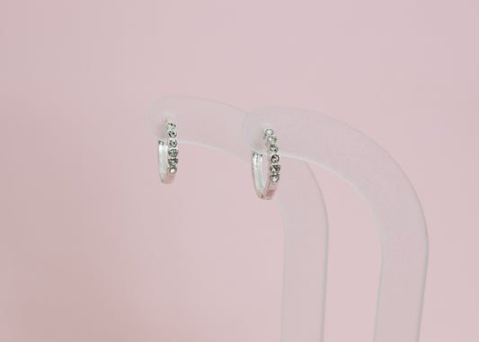 A pair of kids hypoallergenic silver earrings with small clear stones, displayed on a clear acryllic stand with a solid pink background. The earrings are from the brand The Pierced Peanut Earring Co in New Zealand, NZ.