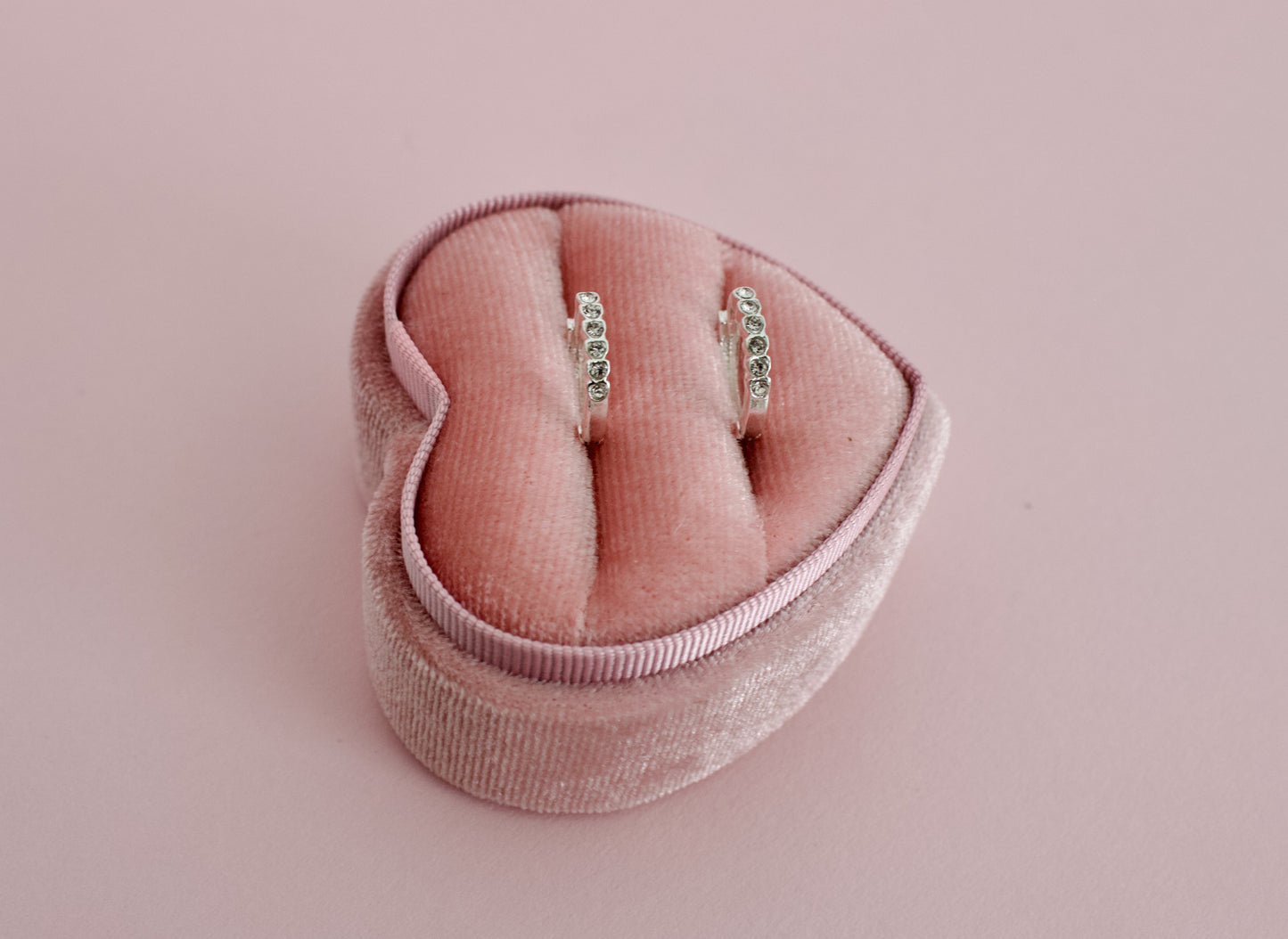 A pair of kids hypoallergenic silver earrings with small clear stones, displayed in a pink velvet heart-shaped ring box on a solid pink background. The earrings are from the brand The Pierced Peanut Earring Co in New Zealand, NZ.