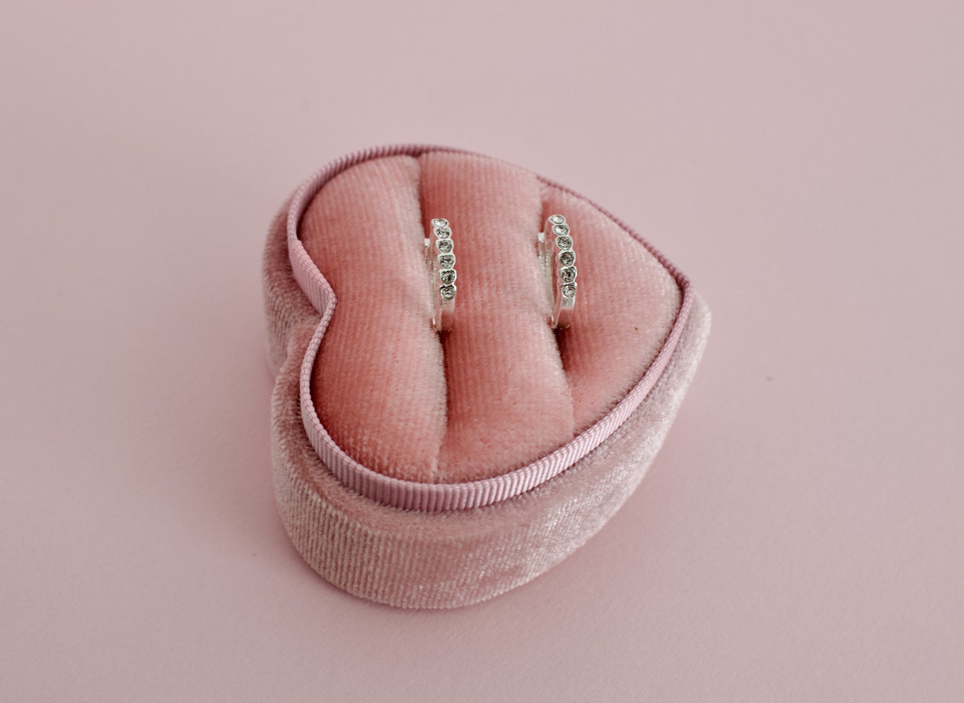 A pair of kids hypoallergenic silver earrings with small clear stones, displayed in a pink velvet heart-shaped ring box on a solid pink background. The earrings are from the brand The Pierced Peanut Earring Co in New Zealand, NZ.